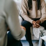 Unrecognizable African American male patient with clasped hands sitting on sofa near blurred anonymous therapist during visit in psychotherapy office